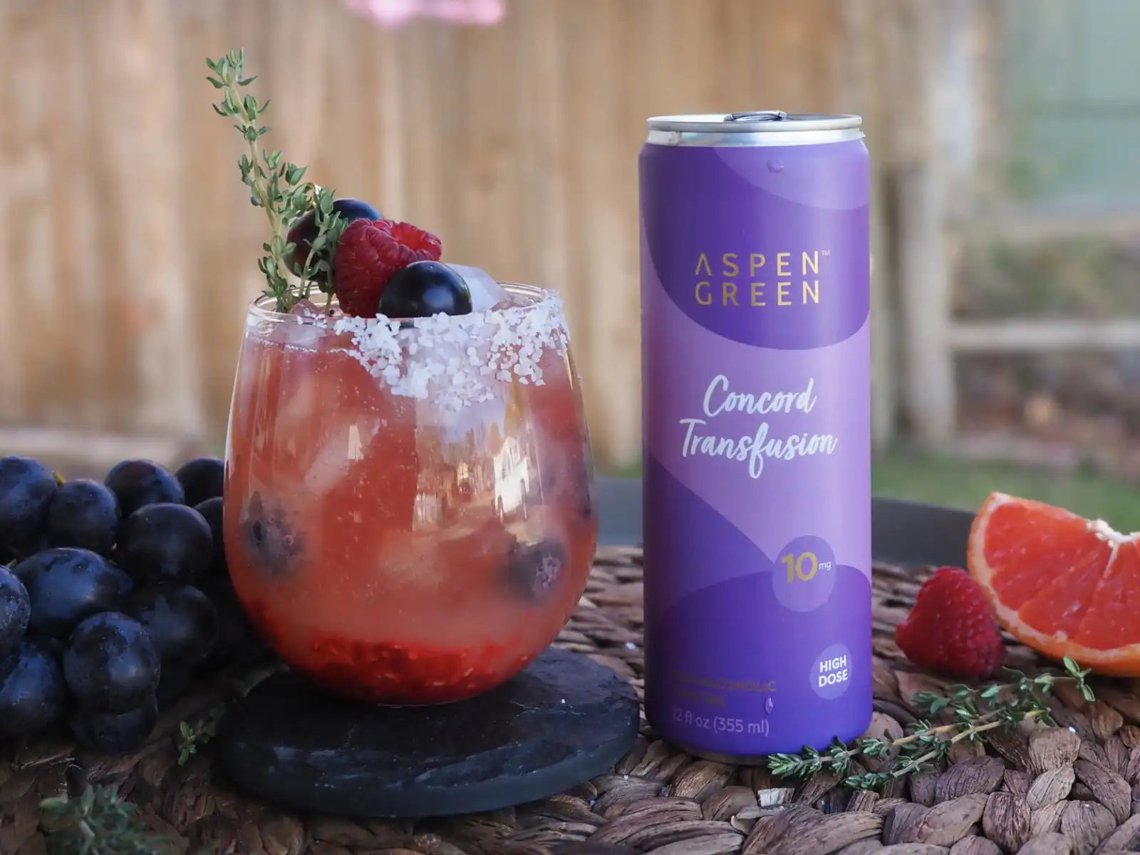 Glass of fruit cocktail with berries and a can of Aspen Green Concord Transfusion on a woven surface.